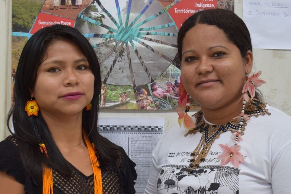 Fotografia de duas mulheres indígenas em frente a um painel com imagens e informações sobre povos indígenas. À esquerda, uma mulher usa blusa preta e acessórios artesanais coloridos. À direita, uma mulher veste camiseta branca e colares e adornos indígenas. Ambas estão de frente para a câmera.